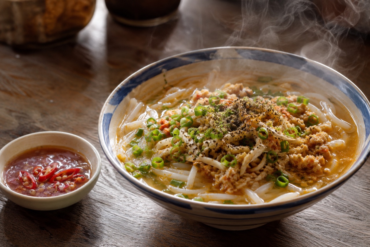 Chén bánh canh sợi trắng ngắn trong nước sánh màu gạch, phủ nhân tôm thịt, hành lá xắt nhỏ và tiêu xay, bên cạnh là chén ớt dằm