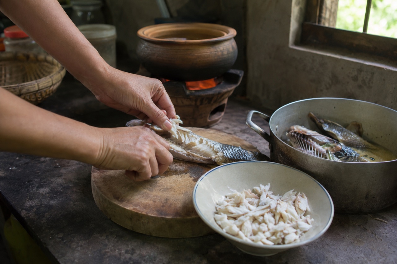 Hai bàn tay gỡ nạc con cá lóc luộc trên thớt gỗ, chén nạc cá trắng để bên cạnh, nồi cá còn nguyên con đặt gần bếp