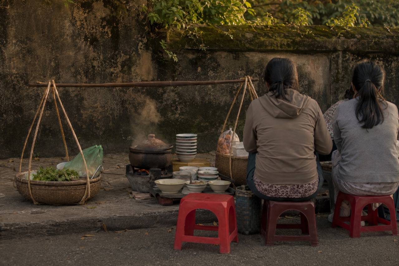 Đôi quang gánh bánh canh dọn xuống lề đường bên bức tường rêu, nồi đất đặt trên bếp than, chồng chén và mấy chiếc ghế nhựa đỏ