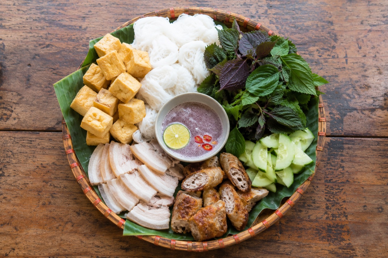 Mẹt tre lót lá chuối bày bún lá cắt khoanh, đậu chiên vàng, thịt luộc, chả chiên, rổ rau kinh giới tía tô và chén mắm tôm đánh bọt