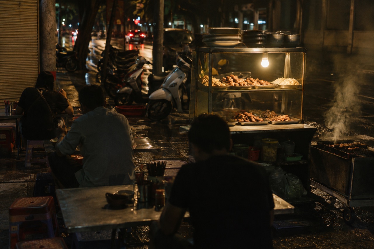 Quán cơm tấm vỉa hè Sài Gòn lúc đêm khuya, tủ kính thắp bóng đèn vàng, khách ngồi ghế nhựa, lò than còn bốc khói bên mép đường