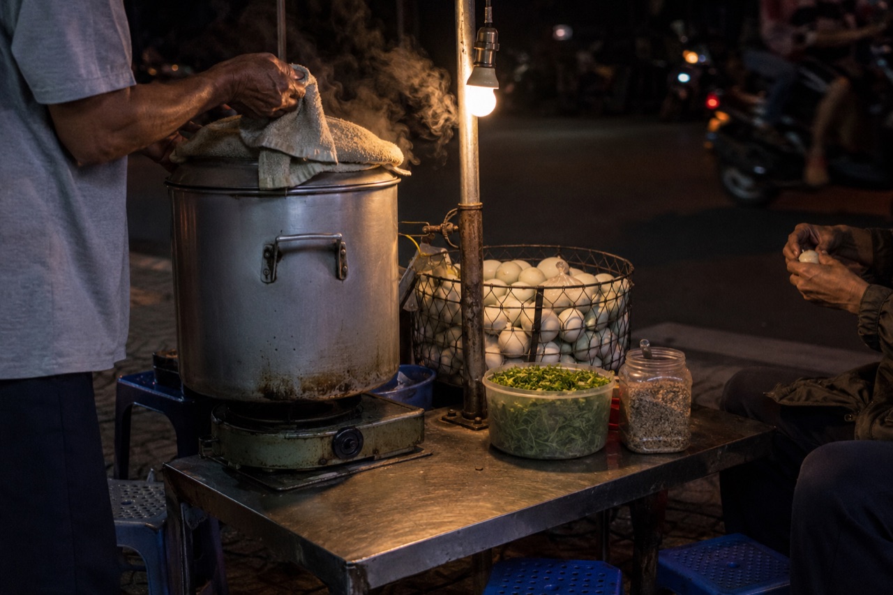 Xe đẩy bán hột vịt lộn ban đêm trên vỉa hè Sài Gòn, nồi nhôm trùm khăn đặt trên bếp, rổ hột, thau rau răm và mấy cái ghế nhựa thấp