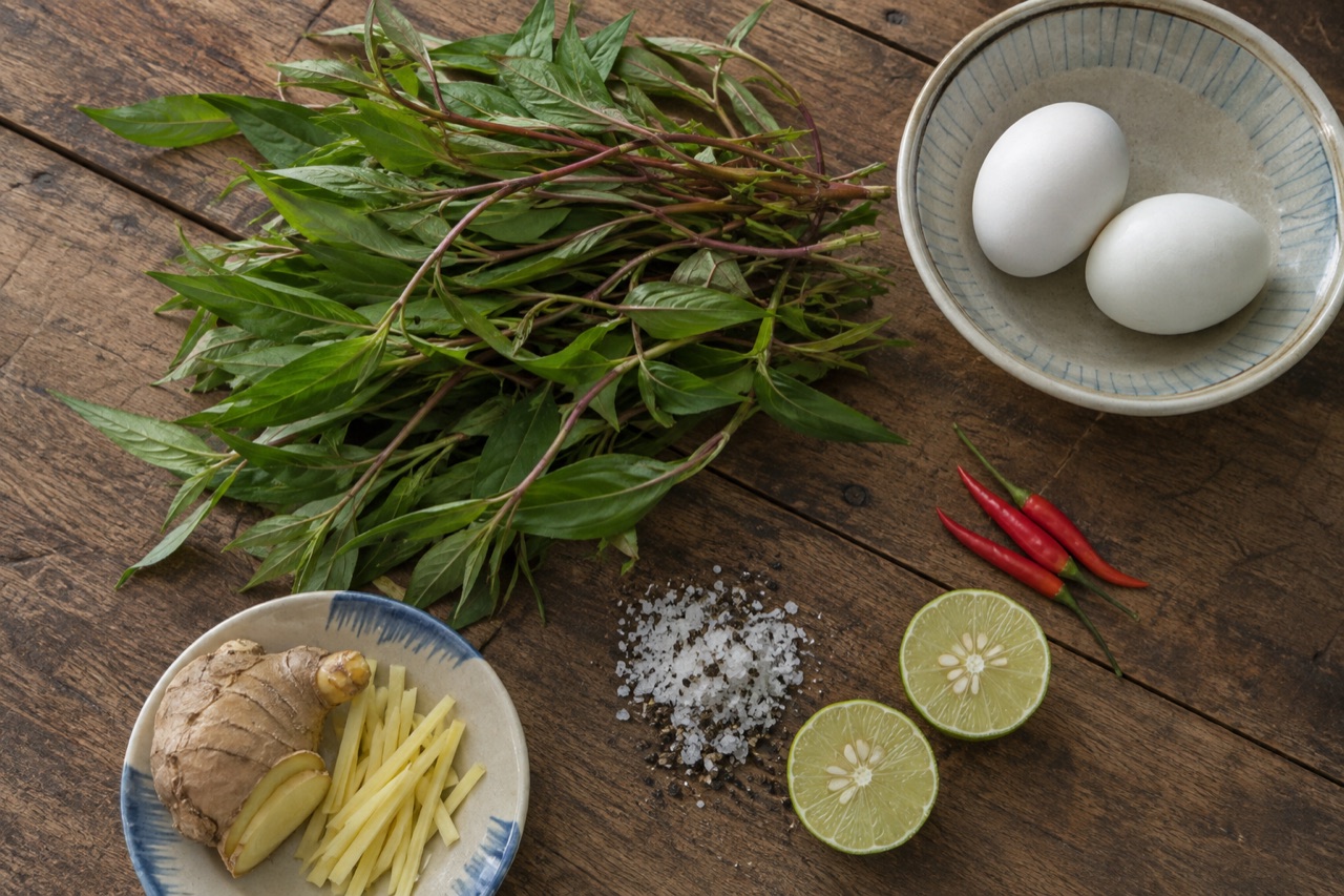Bó rau răm lá nhọn thân đỏ tía trải trên bàn gỗ, kế bên là muối hột trộn tiêu, hai nửa trái chanh, mấy trái ớt hiểm và gừng xắt sợi