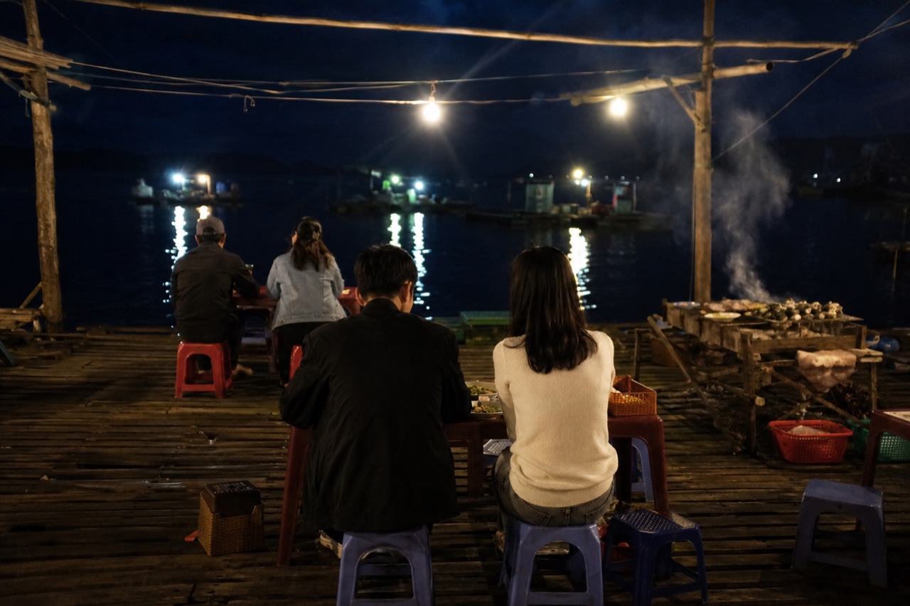 Quán bè trên mặt biển ban đêm, bóng đèn tròn treo trên đầu, bàn ghế nhựa kê trên sàn ván, xa xa là đèn thuyền