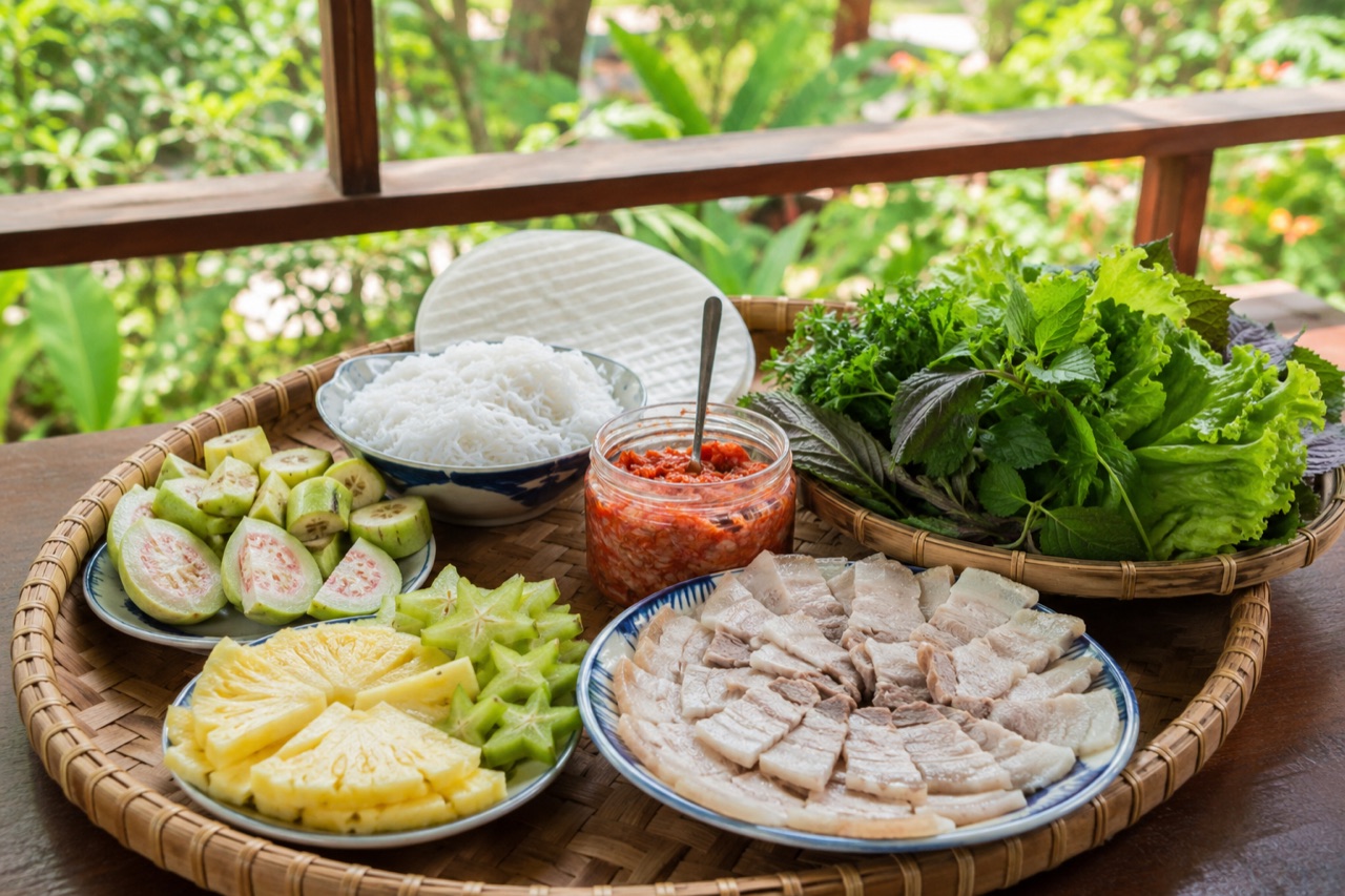 Mâm ăn kèm tôm chua Huế dọn ngoài hiên: dĩa ba chỉ luộc xắt mỏng, rổ rau sống, bún, bánh tráng, khế, thơm và trái vả xắt lát
