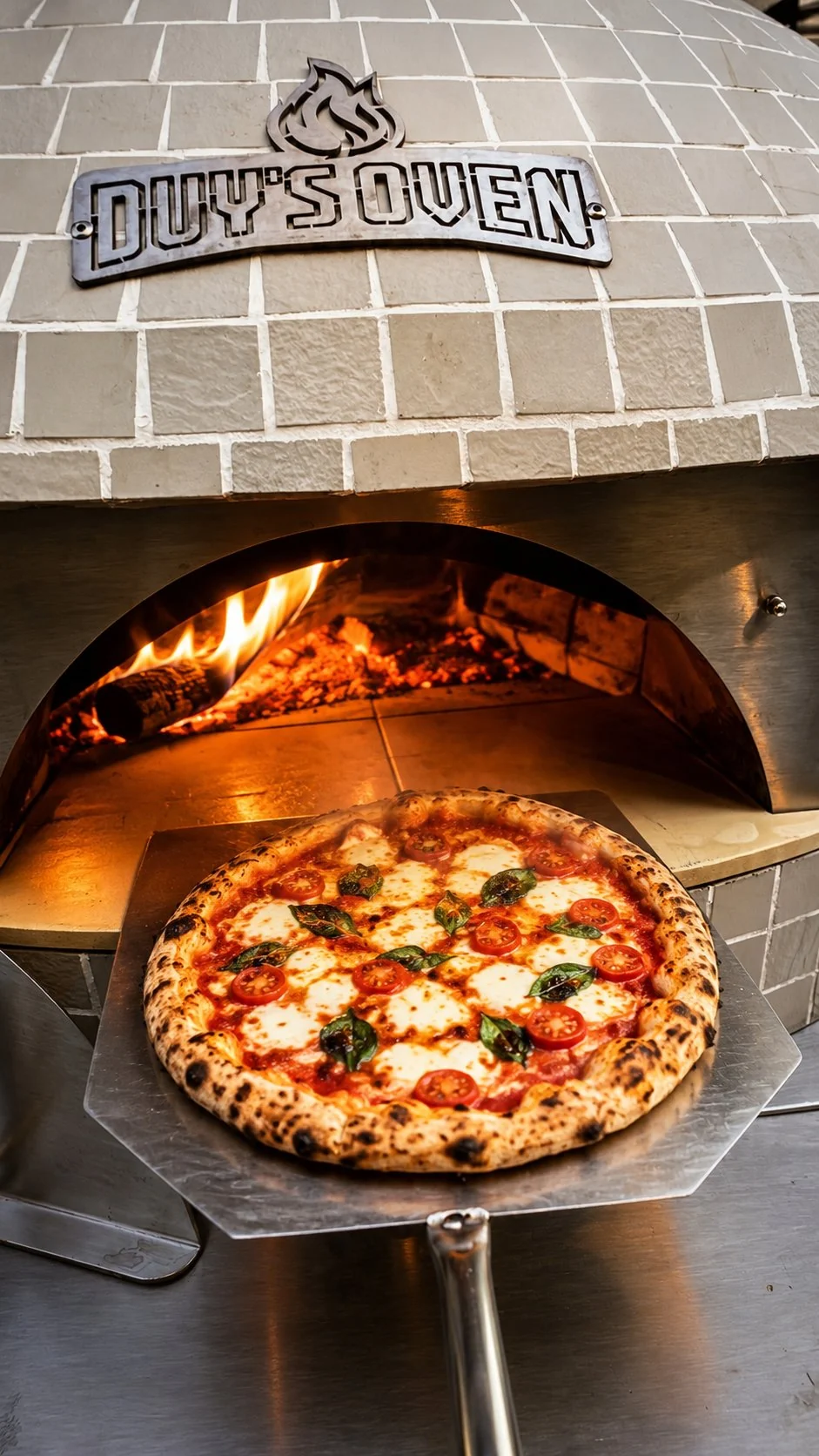 Pizza Margherita vừa ra lò trên xẻng, phía sau là miệng lò đang cháy củi